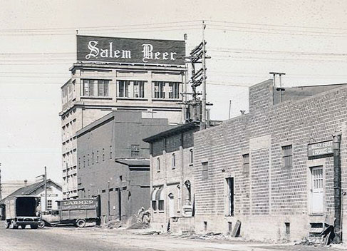 Brick building with sign on top "Salem Beer." Trucks from the 1930s shown on street.