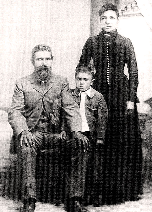 A bearded man sits in a chair with his son and wife standing to his left.