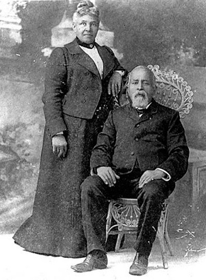 Richard Bogle sits in a wicker chair with his wife, America standing to his right.