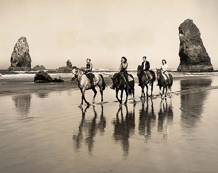 Four people on horseback ride along Cannon beach. Pinnacles of rock stand in the background.