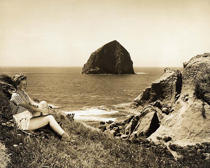 Woman, sitting on a grassy hillside, leans back to admire view of Haystack Rock