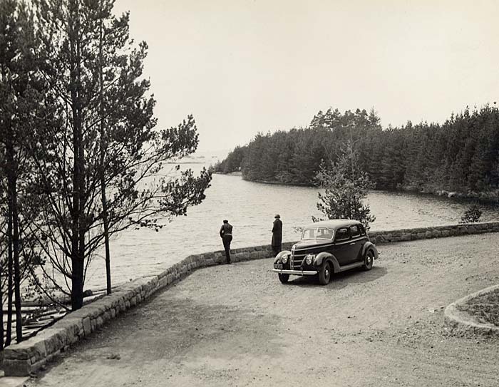 Two people stand outside a car in a turn-around in Honeyman Park overlooking Cleawox Lake.