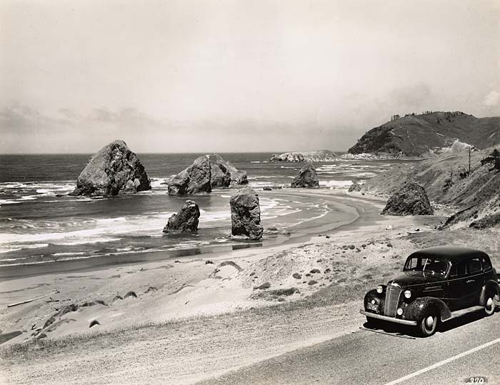 1940 era car drives along highway 101. The ocean is on the left with large rocks jutting out.
