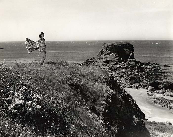 Woman stands on grass covered rock outcrop above the beach. Her wrap flys behind her in the wind. 