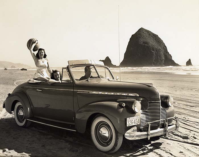 A man drives a Chevrolet along the beach in front of Haystack Rock. Two women ride with him. 