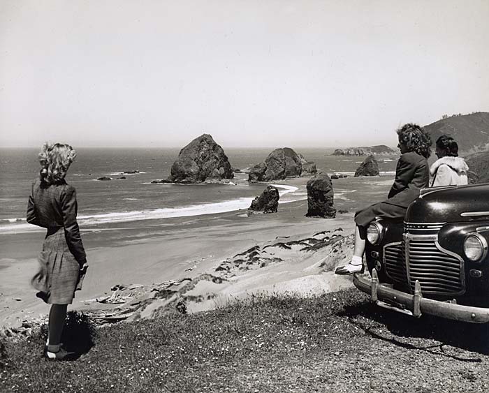 Three women, 1 standing, 2 leaning on a car, look out over the beach.