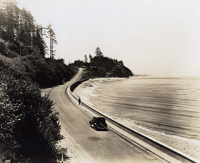 Single car drives along a road on the sea wall. Ocean on right and tall evergreen trees on left.
