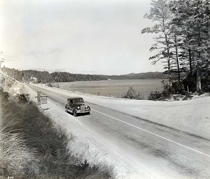 A single car drives along a two lane road with the lake to the right.