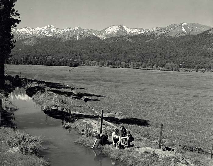 Three people squat near an irrigation ditch. The Elkhorn Mountains are in the background.