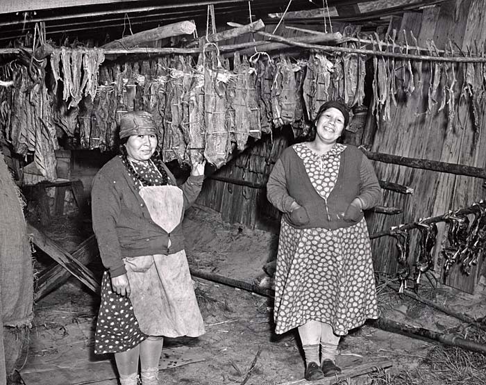 Two Native American women in dresses stand in a fish drying room and smile for camera.