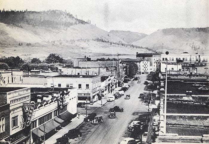 View from above of the main road in La Grande in 1940. Old cars drive on a road lined with businesses. Rolling hills in the back