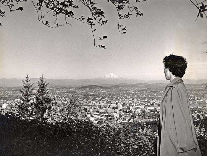 Woman stands to the right looking over Portland from above. Mt. Hood's snowy peek shows in the distance.
