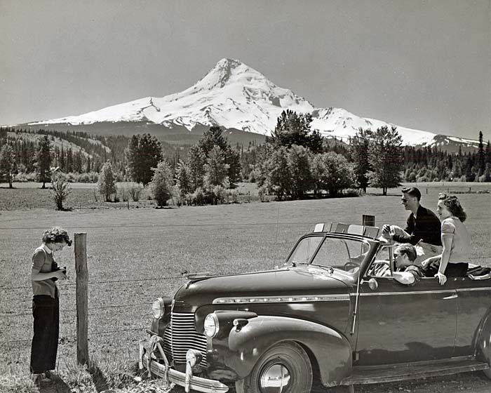 Three young adutls pose for a photo in a 1940s car while another young adult takes a photo. Mt. Hood's snowy top shows in back.