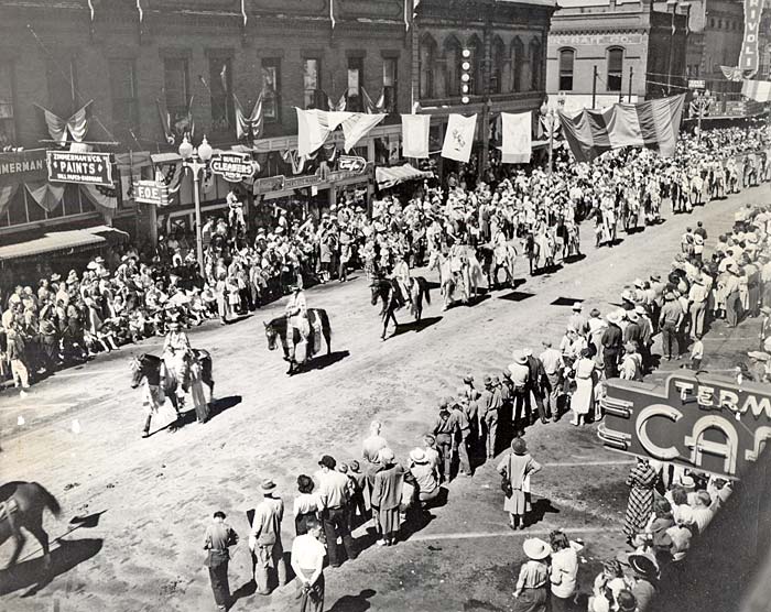 A single file line of a dozen or more horses with riders streams down the middle of a street lined with spectators.