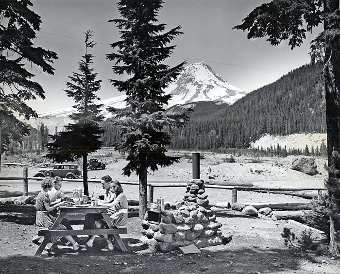 Four young adults, 2 women, 2 men, sit at a picnic table eating a meal. Mt Hood's snowy peek shows in the background.
