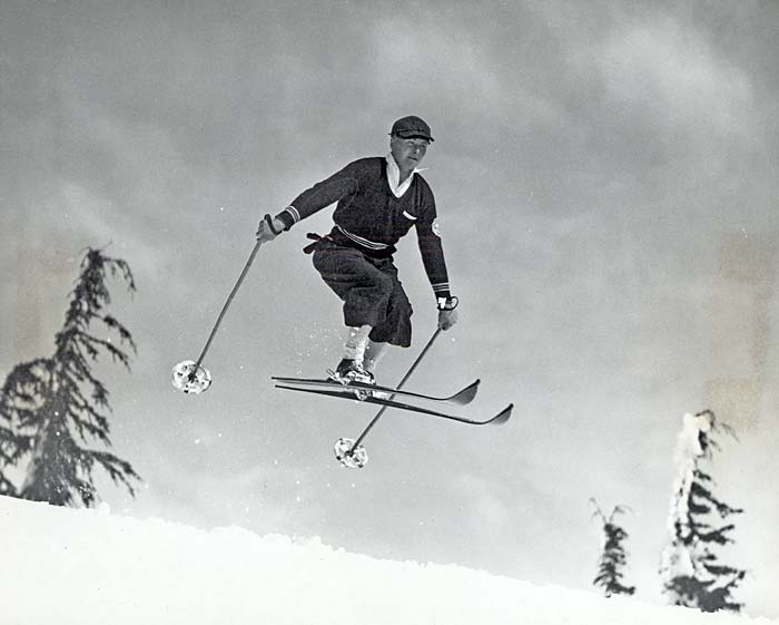 Skier in mid jump over a snowy hilltop. The tops of evergreen trees are seen in the background.