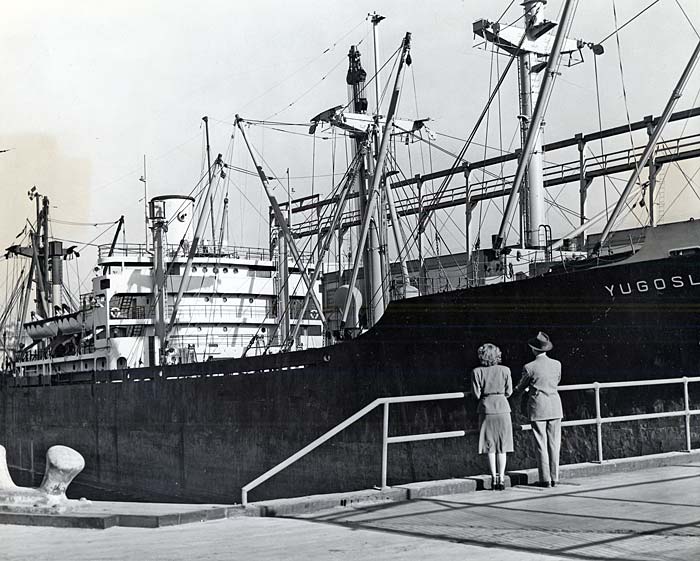 A man and a woman stand on the sidewalk holding on to a railing and looking at a large ship.