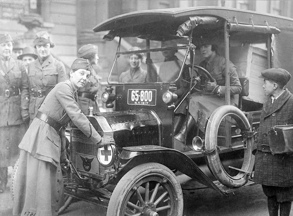 Photo of 2 women sitting in a car while another woman stands at the front of the car ready to crank the engine.