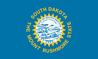South Dakota flag has the state seal on a blazing sun in the center against a blue background. South Dakota is written above the seal; The Mount Rushmore State written below.
