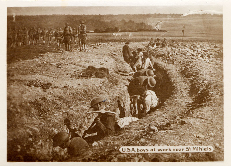 A dozen men in a trench with pick axes work while other men above look on.