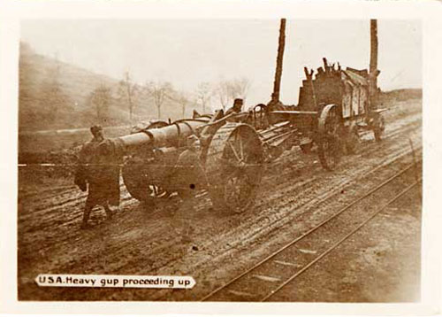A truck pulls artillery gun up a hill on a dirt road with two men at the rear.