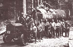 Log truck with about 16 men gathered around stands in the forest loaded with logs.