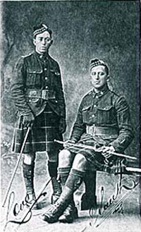 Two men in kilts, one man standing, one sitting, pose for picture.