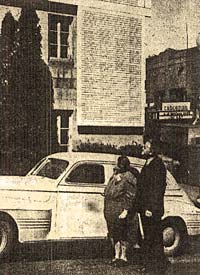 Woman & man stand next to a car outside the county courthouse and look at the honor roll on the wall.