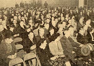 Hundreds of people sit in a crowded room in folding chairs.