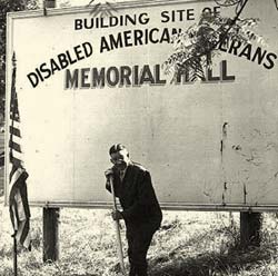 Gov. McKay with a shovel posing in front of sign that reads "Building site of disabled American Veterans memorial hall"