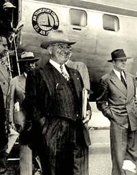 President Truman in suit and cowboy hat stands with other men outside an airplane.