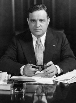 Photo of Fiorello La Guardia in suit and tie sitting at desk with papers and books in front of him. He holds eye glasses.