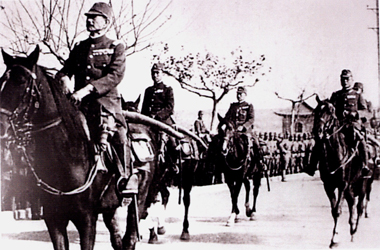 Japanese men on horses ride past lines of soldiers.