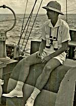 Man in pith hat, shorts, t-shirt sits on a boat. He has binoculars around his neck.