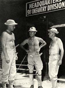 3 shirtless men in khaki pants and boots talk outside a building with a sign "Headquarters 91st Infantry Division"