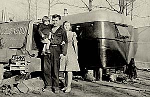 Photo of man holding young child with his wife standing next to him. They stand outside a trailer in a trailer park.