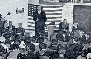 Photo of man on stage by podium in front of American flag talking to a group of dozens of people.