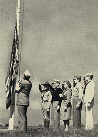 A man in uniform raises an American flag on a pole on a grassy lawn while 5 youths stand at attention.