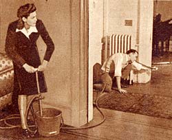 Photo of woman with bucket and pump handle on one side of living room wall. Man holding hose toward something in the other room.