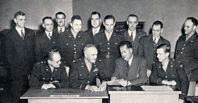 9 men standing, 4 men sitting at a desk examining a document. All dressed in officer's uniforms.
