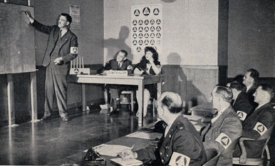 Man stands at front of classroom pointing to something on a map. Class of men and 1 woman look on.