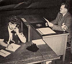 Woman sits at a desk talking on the phone and writing on a paper. Man sits at desk next to her looking at document.
