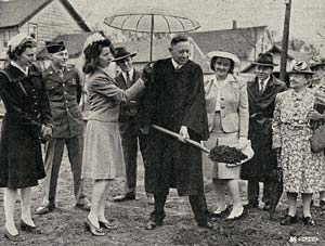 Photo of minister with shovel pulling dirt out of ground. A woman holds an umbrella over him and 6 others stand to watch.