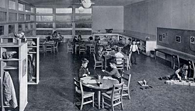 Photo of a classroom with desks, chairs and places to hang coats or store toys. A few children sit at tables or play on floor. 