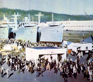 Photo of hundreds of people walking around a shipyard in Portland. 3 naval ships show in the background.