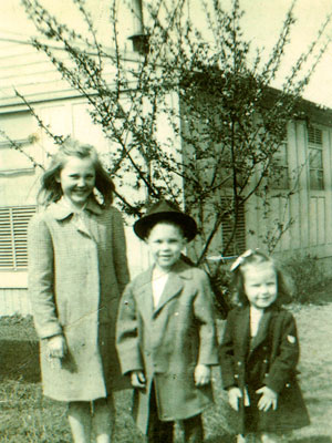 3 children stand in front of a house wearing coats.