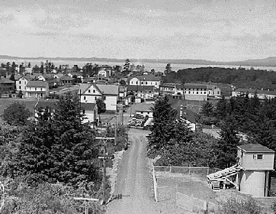 Typical looking community of 1942 with buildings, dirt roads and trees to the side. The ocean in the distance.