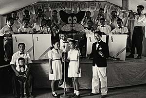 Band sits behind 2 girls singing along with 3 men and a boy. All are dressed in white with ties.