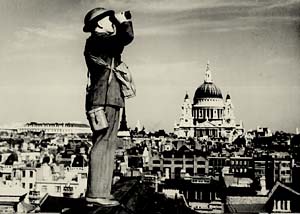 Man in army dress looks through binoculars at sky.