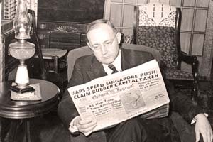 Gov. Sprague sitting in a chair reading a newspaper with the headline "Japs Speed Singapore push claim rubber capital taken"